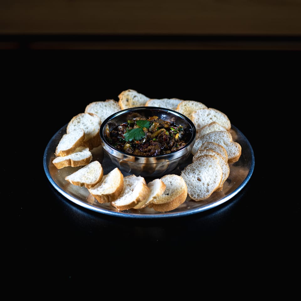 A Vietnamese appetizer of dark, savory pate in a bowl, surrounded by toasted baguette slices on a silver tray.