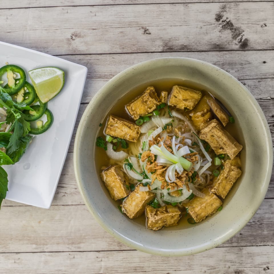 A bowl of Pho Binh's Vietnamese pho with tofu, noodles, and broth, served with a side of jalapeños and lime.