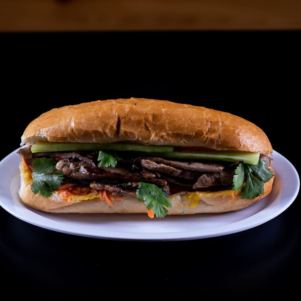 A Vietnamese banh mi sandwich with grilled meat, cilantro, and pickled vegetables on a white plate.